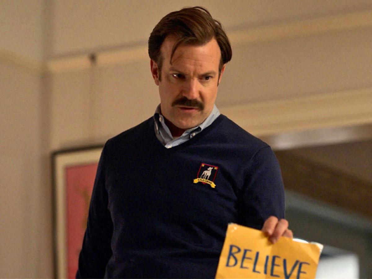 A man in a blue jumper holds a yellow sign reading 'believe'.