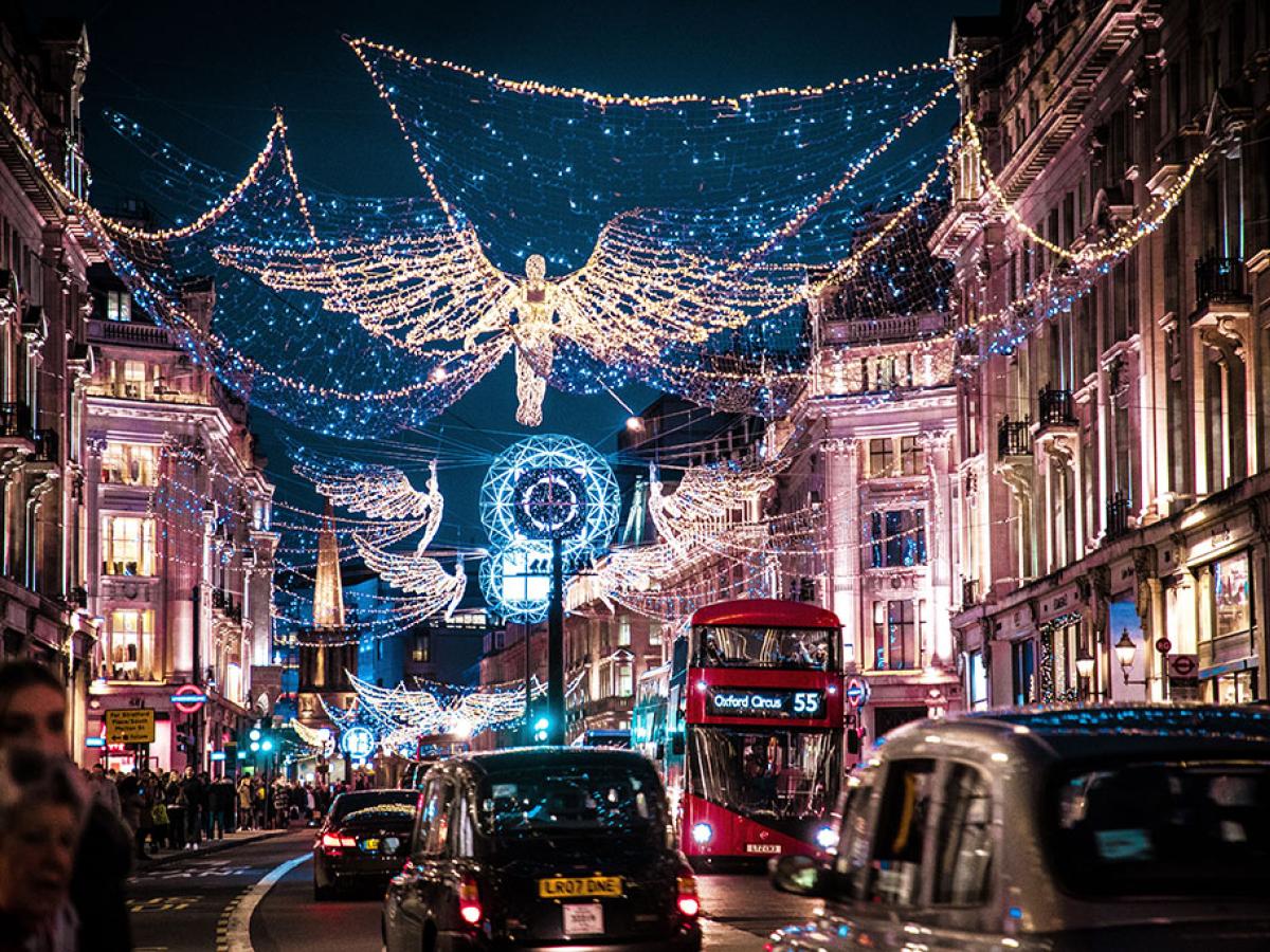 A shopping street is crowded by taxi cabs and buses while above it a Christmas illumination of an angel hangs over all.