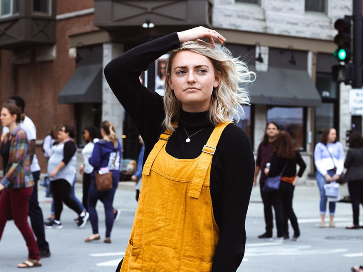 A woman stops in her stride down a street and pensively runs her hand through her hair as she looks to the side.