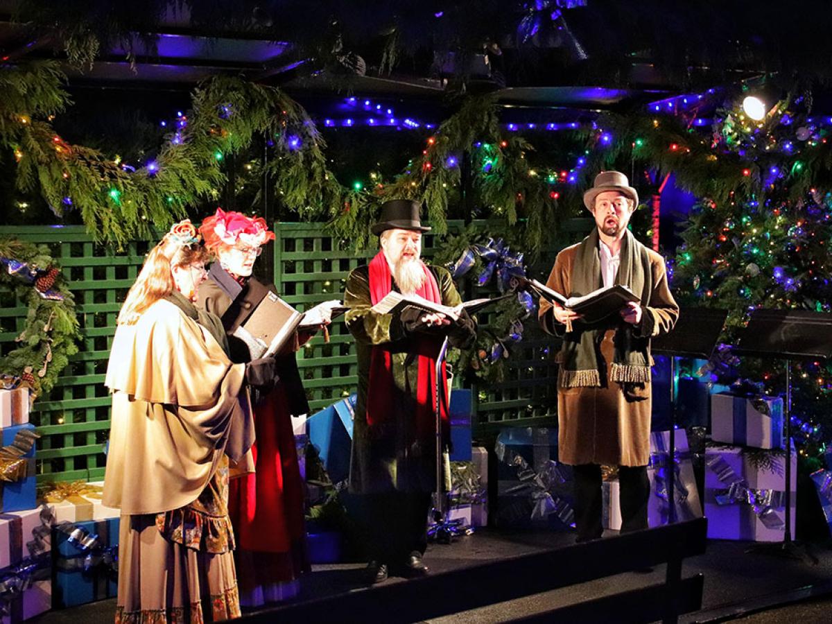 Dressed in Victorian clothes, a group of carol singers stand and sing amid Christmas foliage.