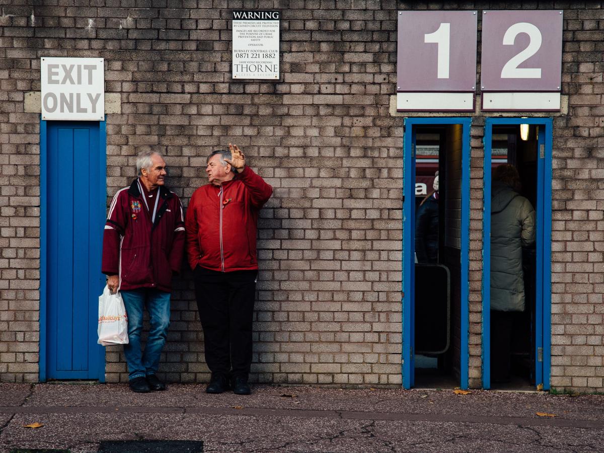 Two older men lean agains the wall of a football club's entrance, next to thin open doorways.