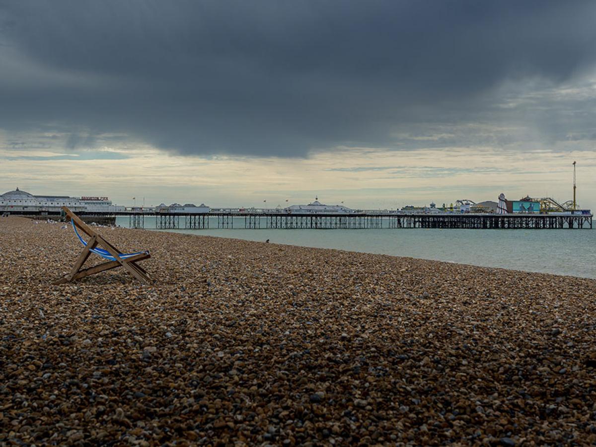 A moody sky overshadows a shingle beach on which a lone empty deckchair stands. A pier with funfair is in the middle distance.