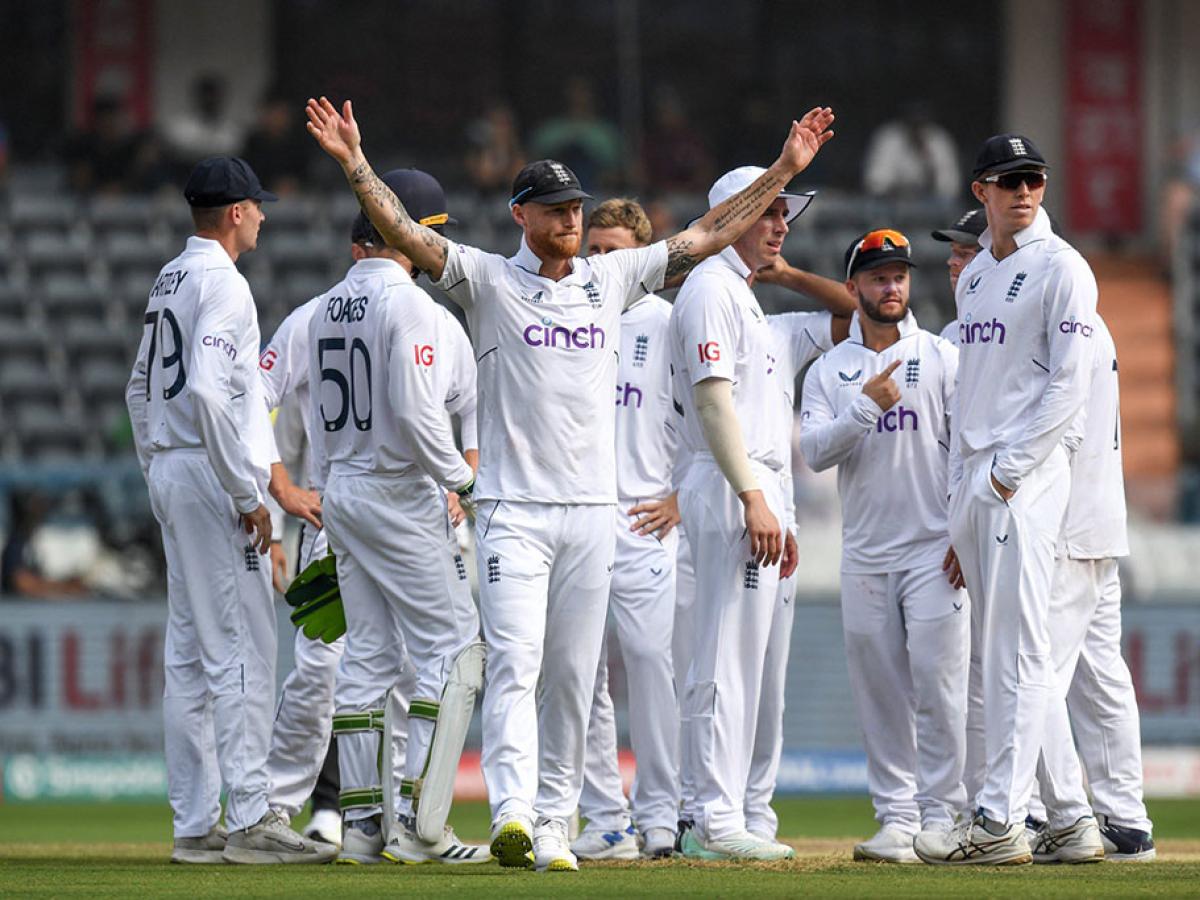 A gaggle of cricket players, dressed in whites, stand on the field. One raises there arm