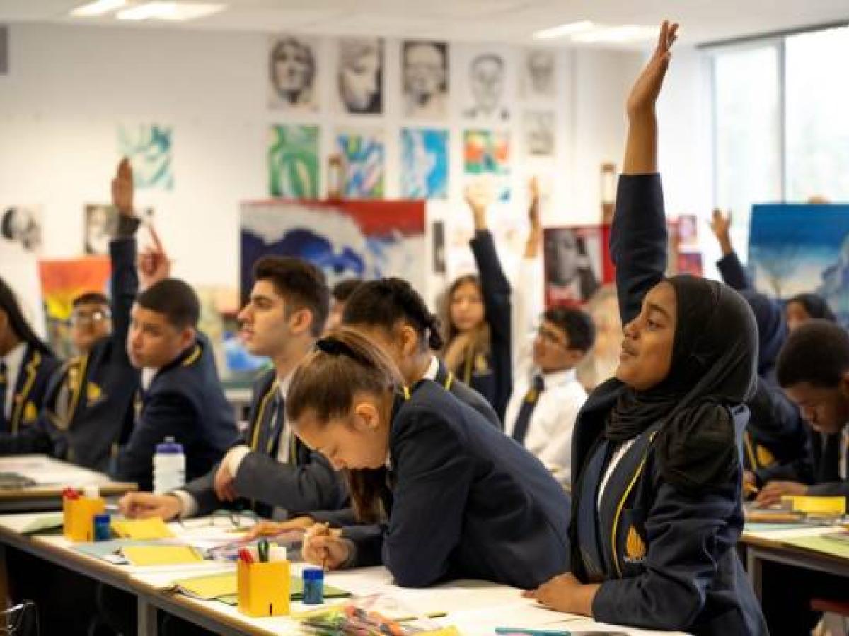 school pupils sit at desk, some with a hand raised.