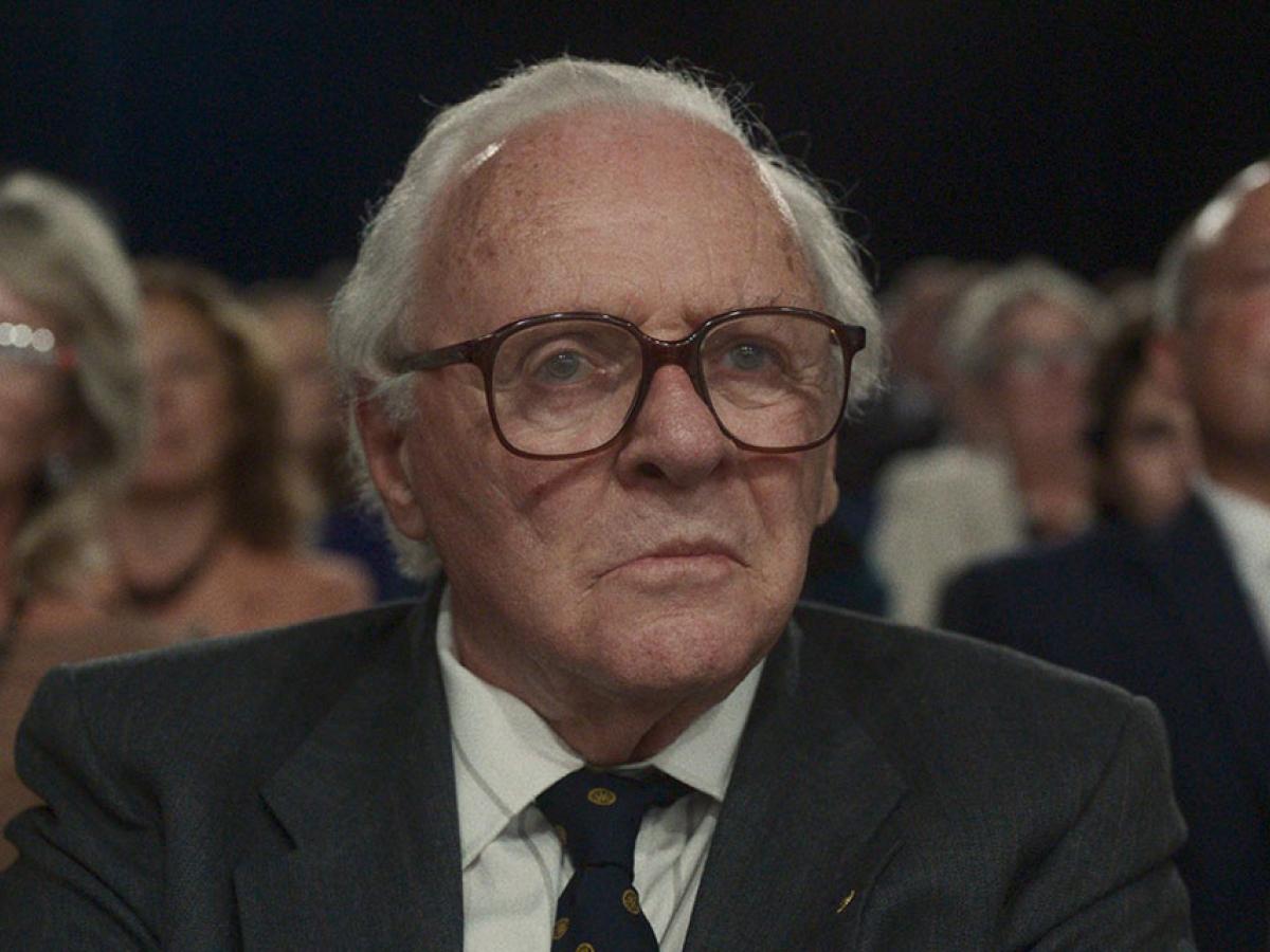 An old man wearing a suit and tie sits in a TV audience as people stand around him.