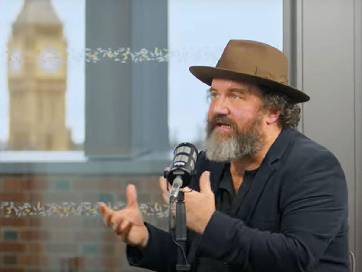 A man wearing a hat sits at a table talking and raises both hands in front of himself to gesture