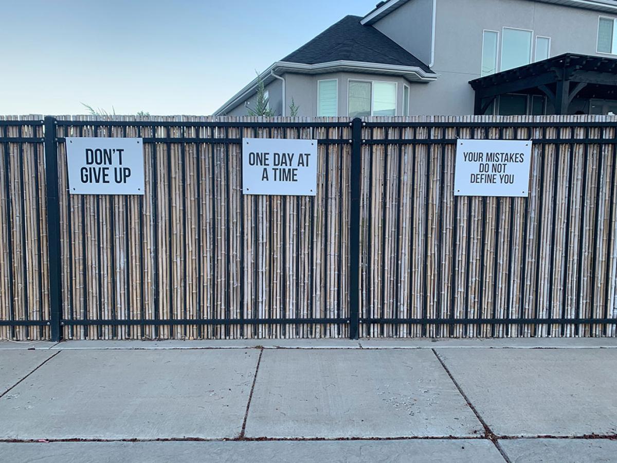 Three signs attached to a fence read: Don't Give Up, One Day At A Time, and Your Mistakes Don't Define You.