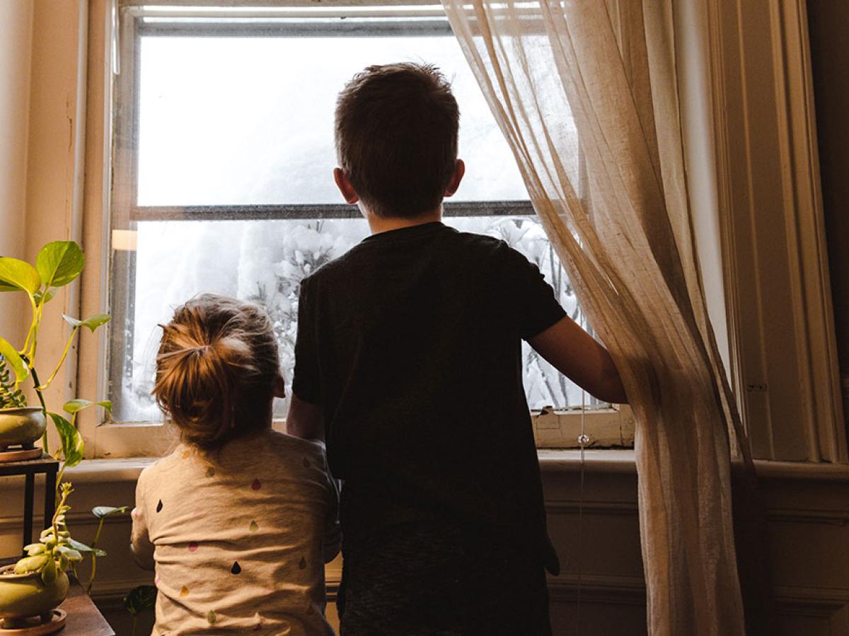 Two small chidlren push a curtain aside to stare out the window at snow.