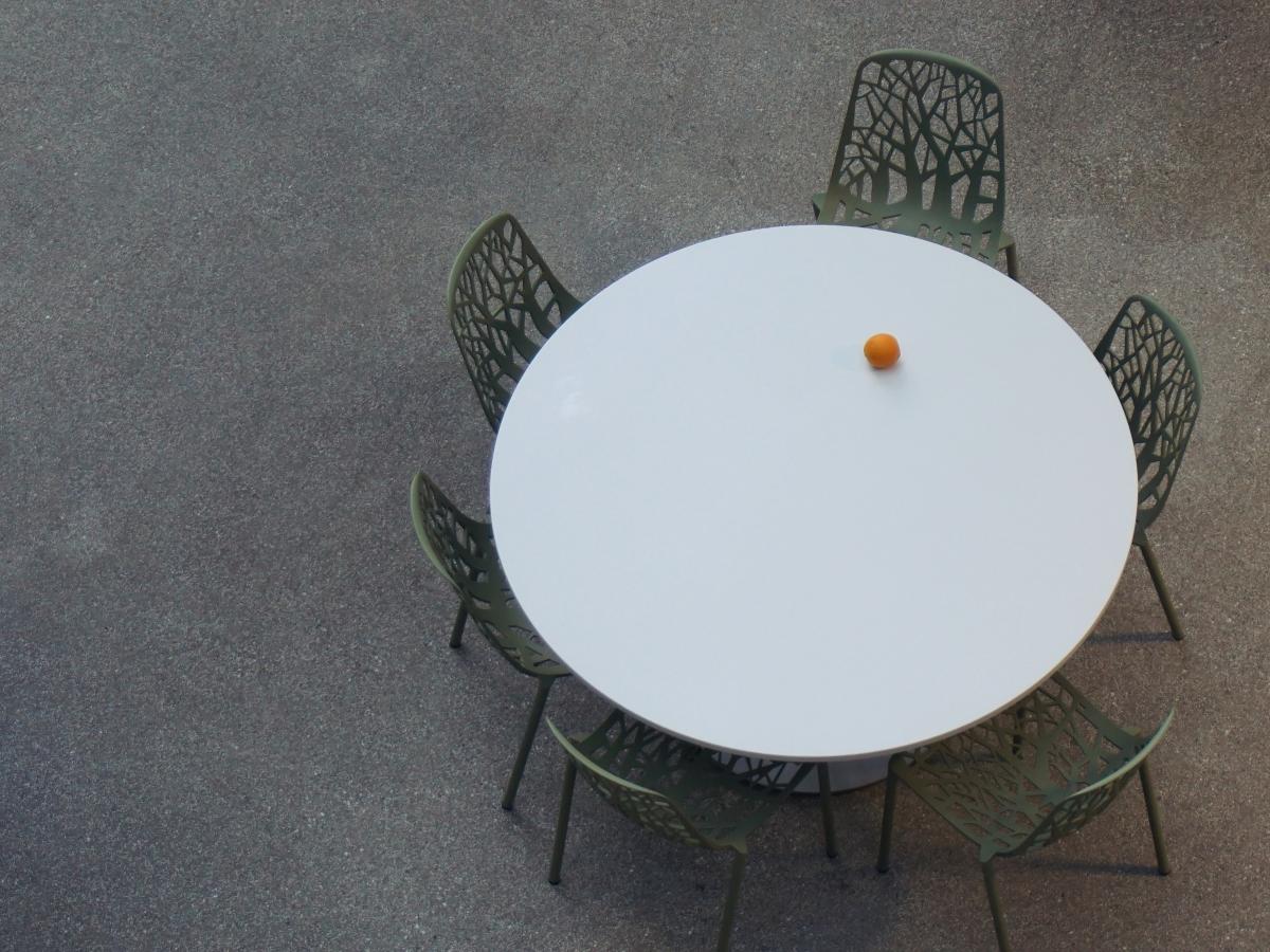 a round table with empty chairs is seen from above. An orange is the only item on it.