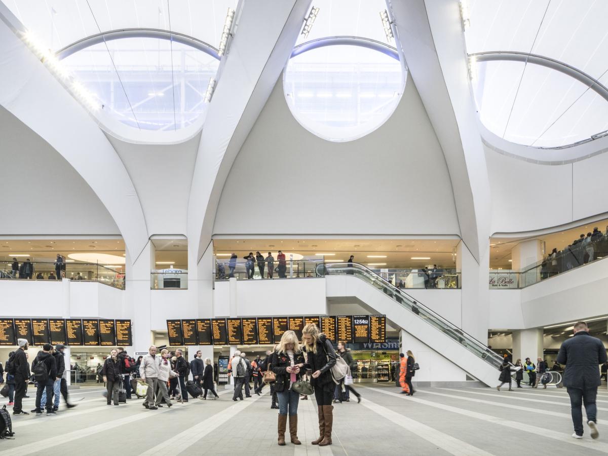 A airy and light station concourse in which people stand and look at a long set of travel information screen.