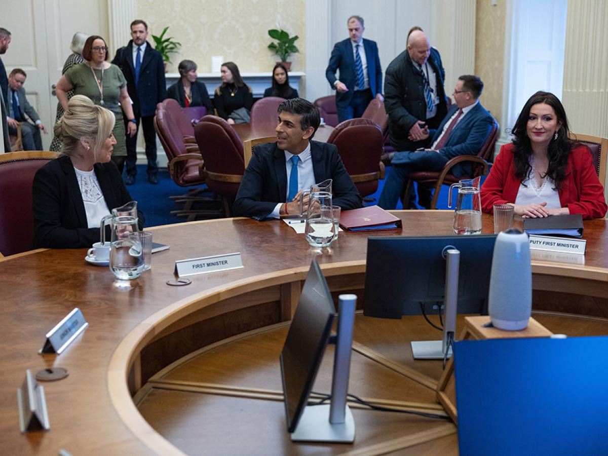 Smartly dressed politicians sit or mill around a round table.