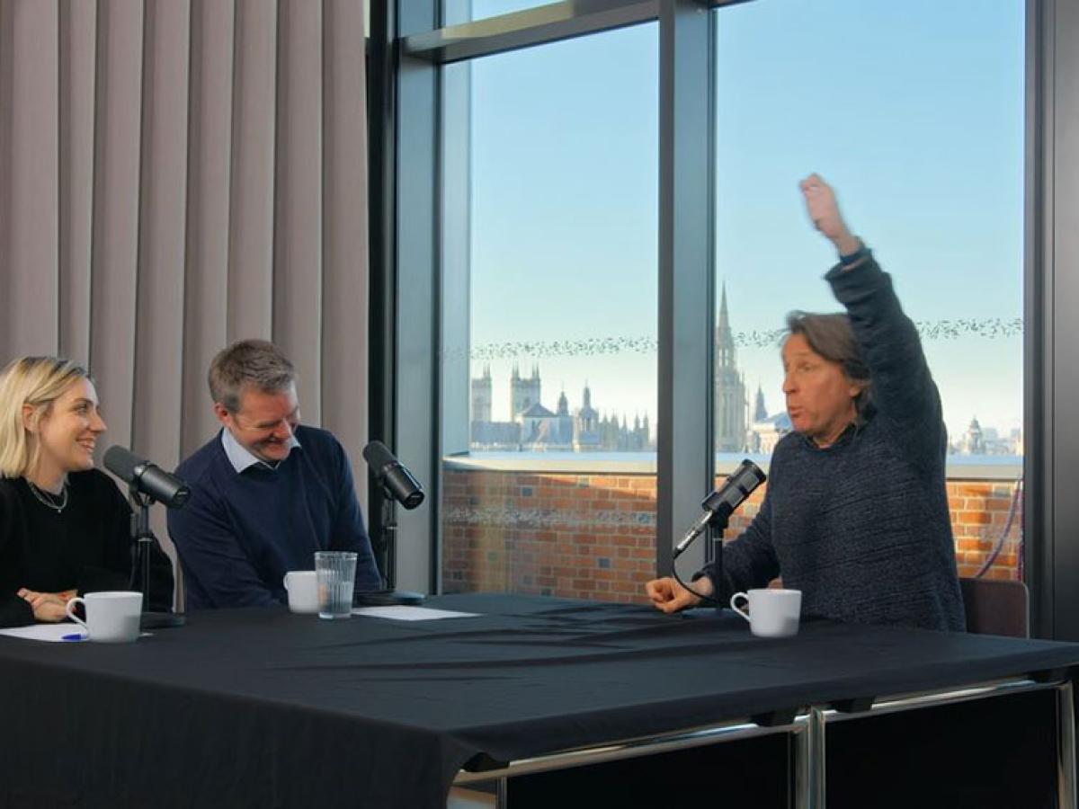 A sitting man raises one hand above his head to emphasise a point, while two other people looking on are laughing.