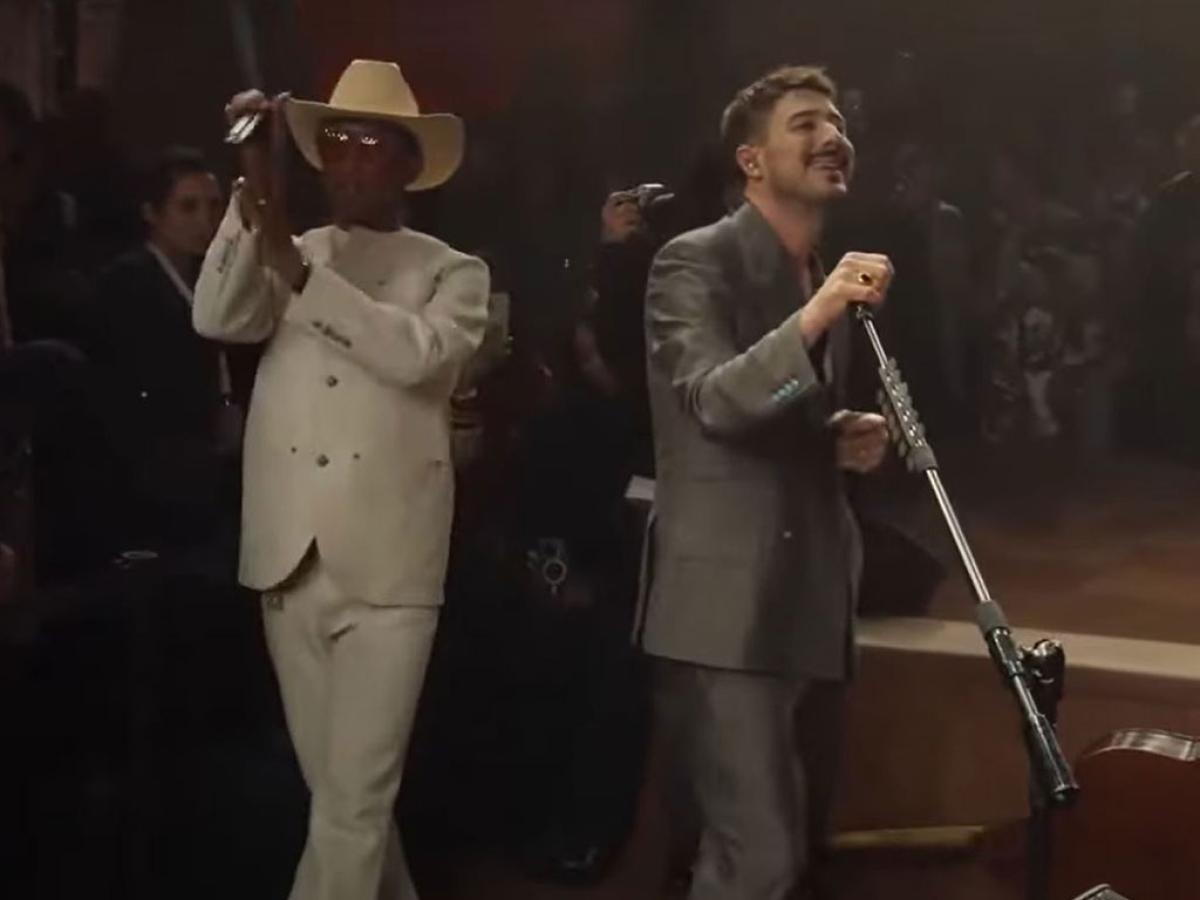 Two singers peform together. One in a white suit and stetson claps their hands. The other tilts the mic stand.