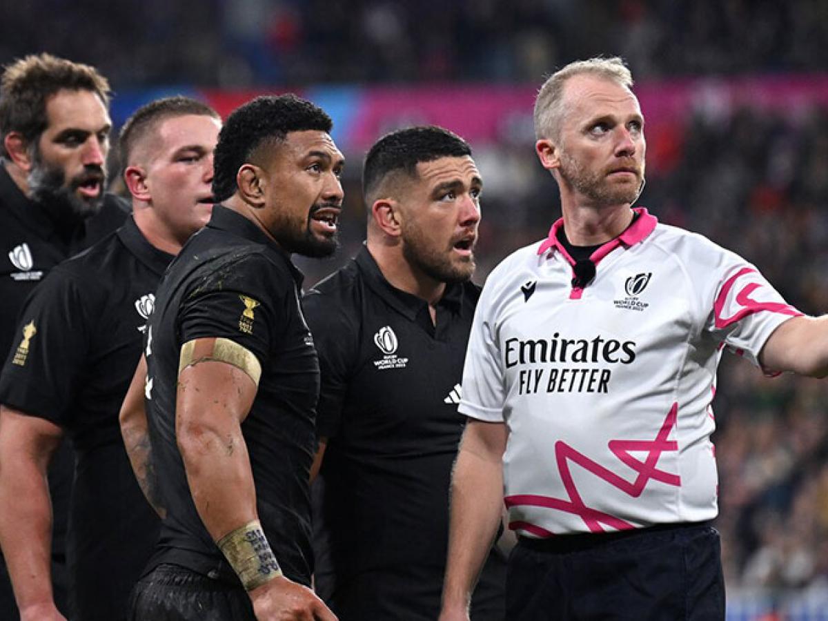 Four rugby players stand and watch beside a referee gesturing with his arm.