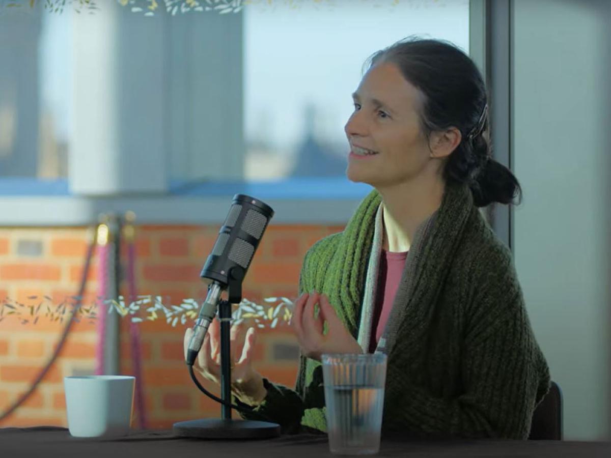 a woman sits at a table, with a mic in front of her, talking and holding her hands out in front of her.