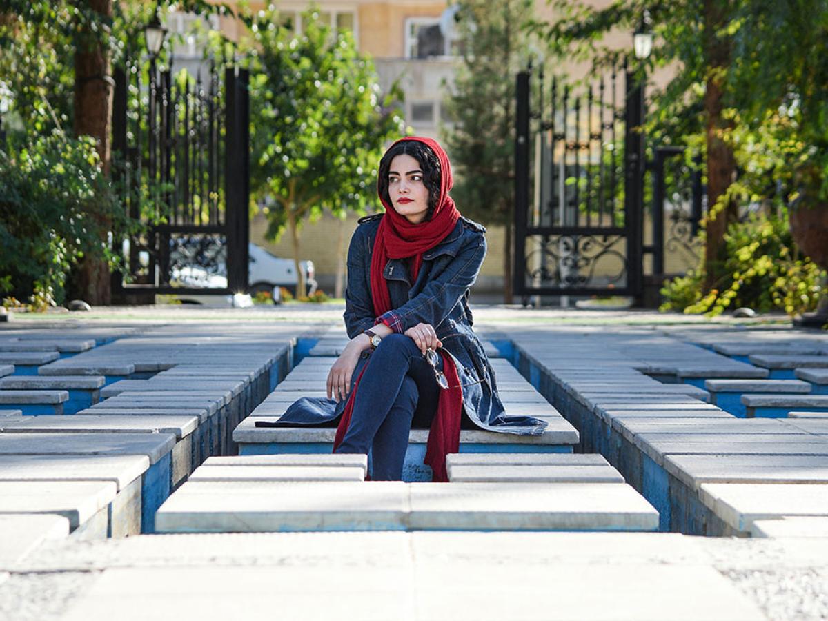 A woman wearing a headscarf sits, looking pensive, amid a grid of concrete seats.