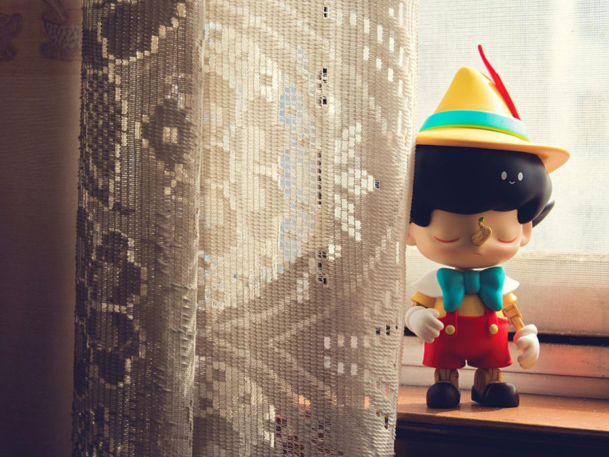 A Pinnochio figure stands on a window sill beside some net curtains.