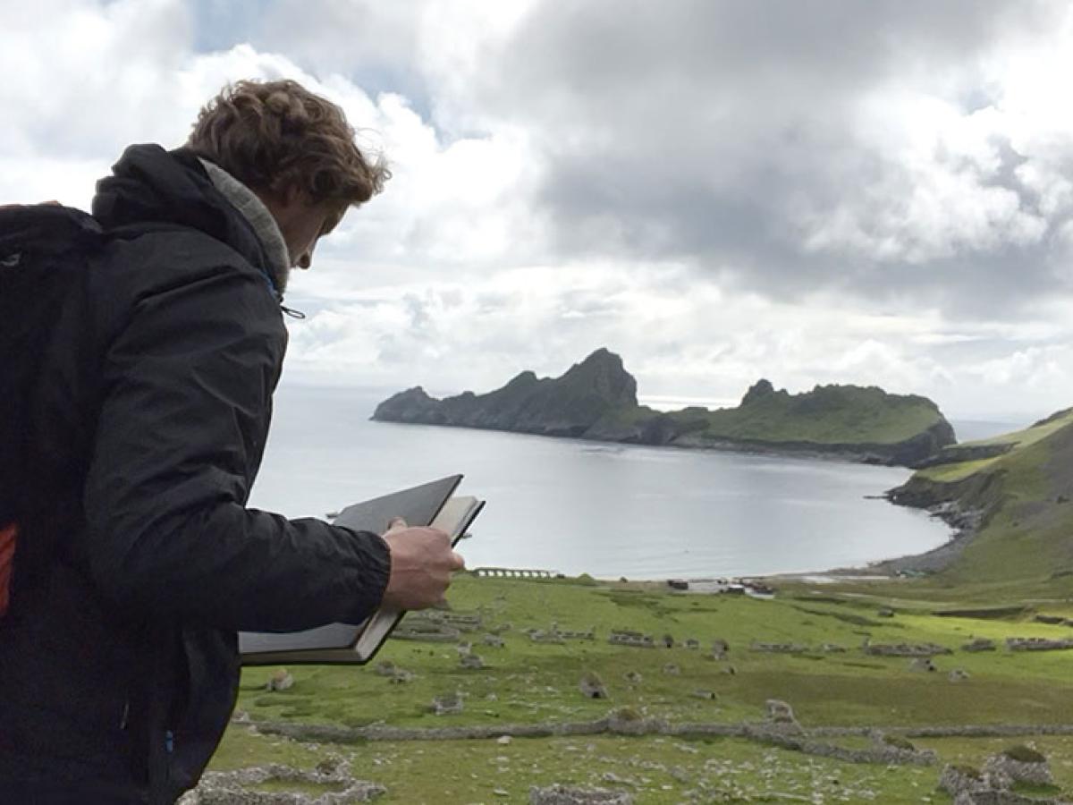 An artist holds a sketchbook while standing overlooking a deserted village by a bay, sided by jagged cliffs.