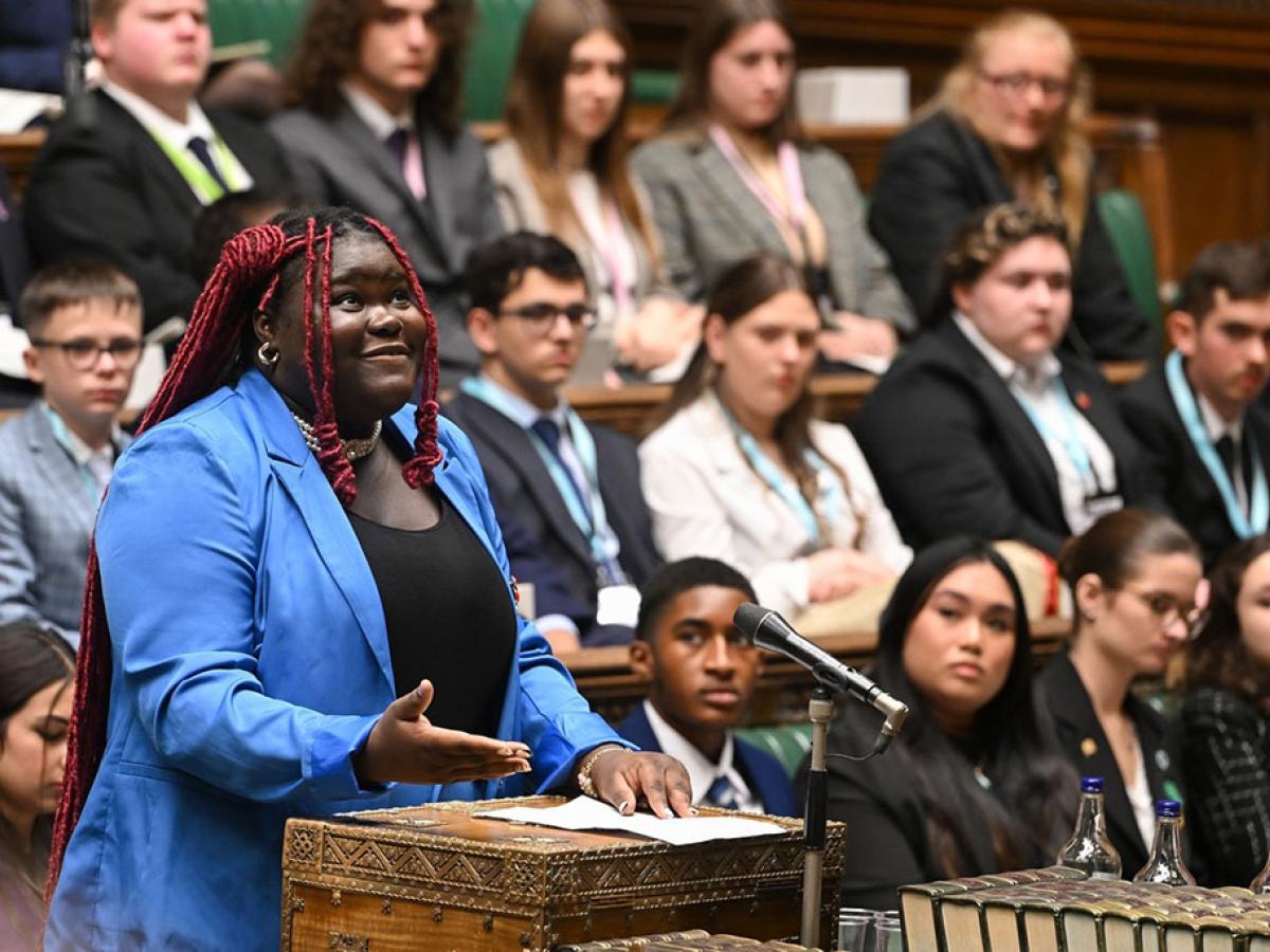 A young woman in a blue suit stands at a wooden box in a parliamentary debating chamber looking upward while speaking. 