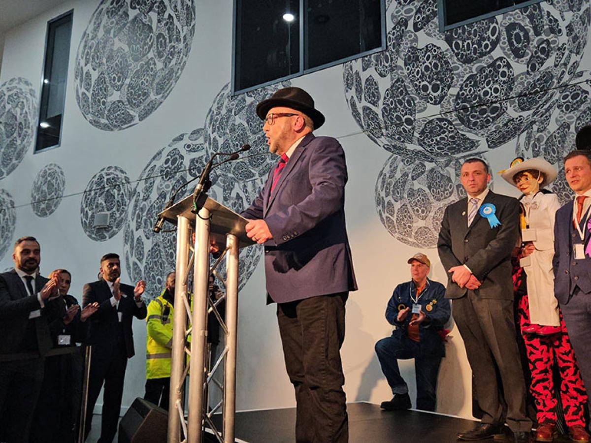 A winning candidate at an election address the audience from a lectern while the loosing candidates look on