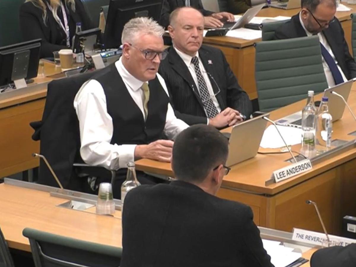 A man wearing a waist coats sits at a desk and ask a questions of a panel of people with their backs facing us.