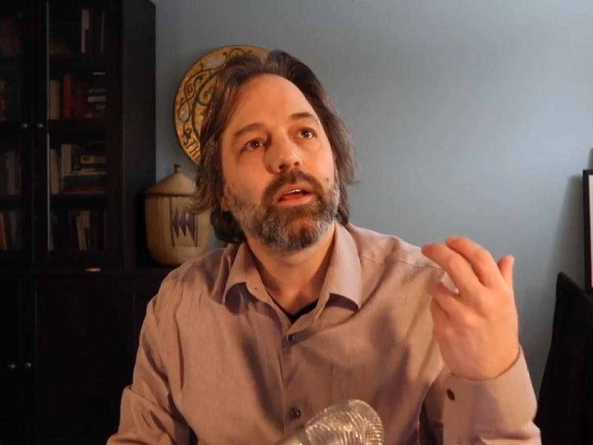 A man sits at a desk and looks up and gestures with his hand.