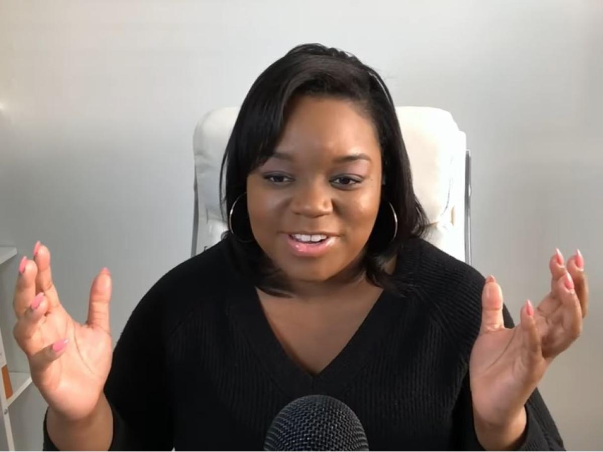 A woman sits and speaks into a desktop microphone and holds both hands up and out expressively.