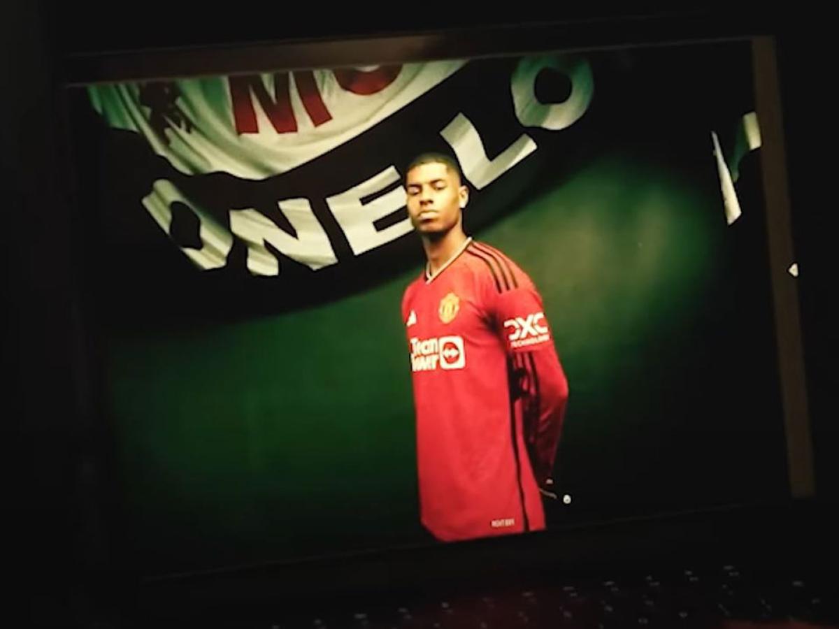 A professional footballer wearign a red top stands proudly beneath a white and black banner.