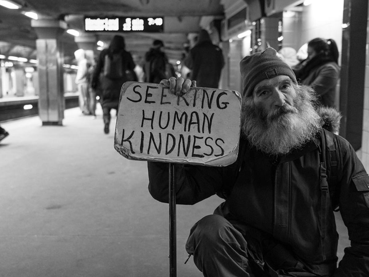 a man in a wheelchair sits in a subway station holding a sign reading 'seeking human kindness'.