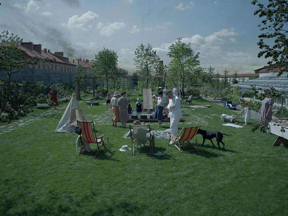 in an immaculate garden a family play in and around a small swimming pool. Beyond the garden wall, a barracks is visble with crematorium smoke rising beyond it.