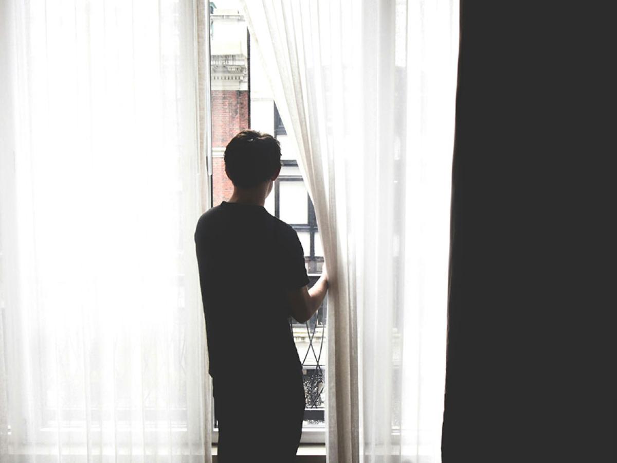 A persons stands, holding a net curtain aside to gaze out.