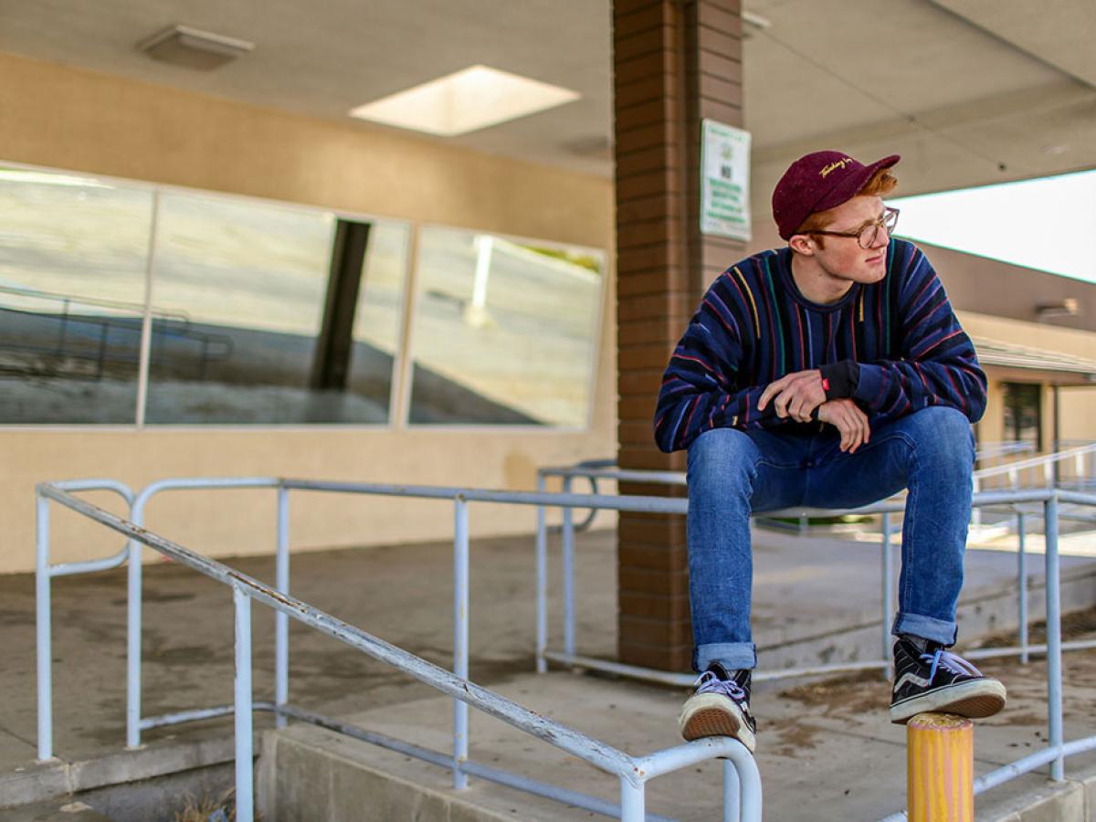 A casually dressed man perches on railing balancing, clasping his hands and looking around.