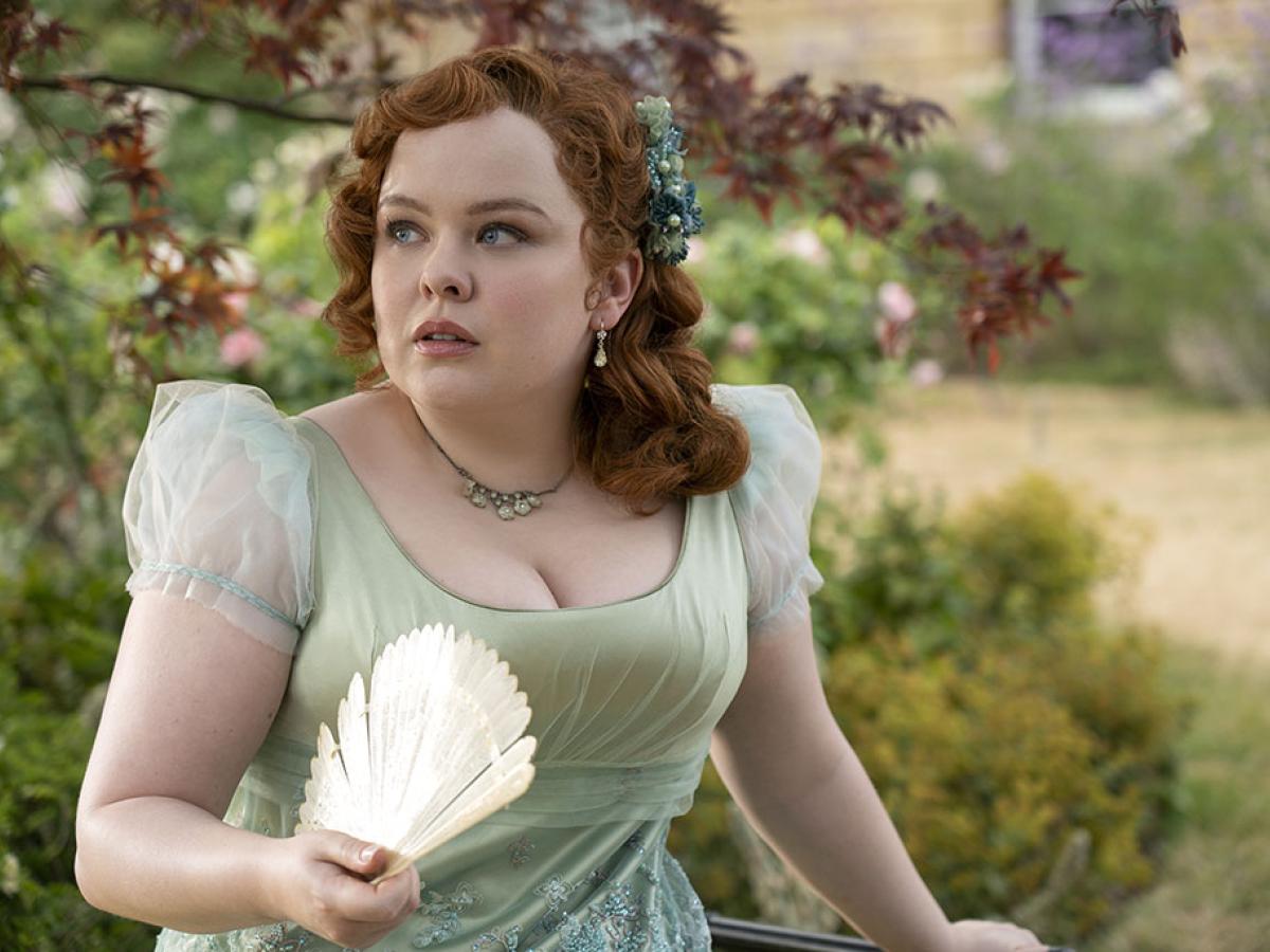 A young lady in Regency dress, holds a fan while looking around a garden.