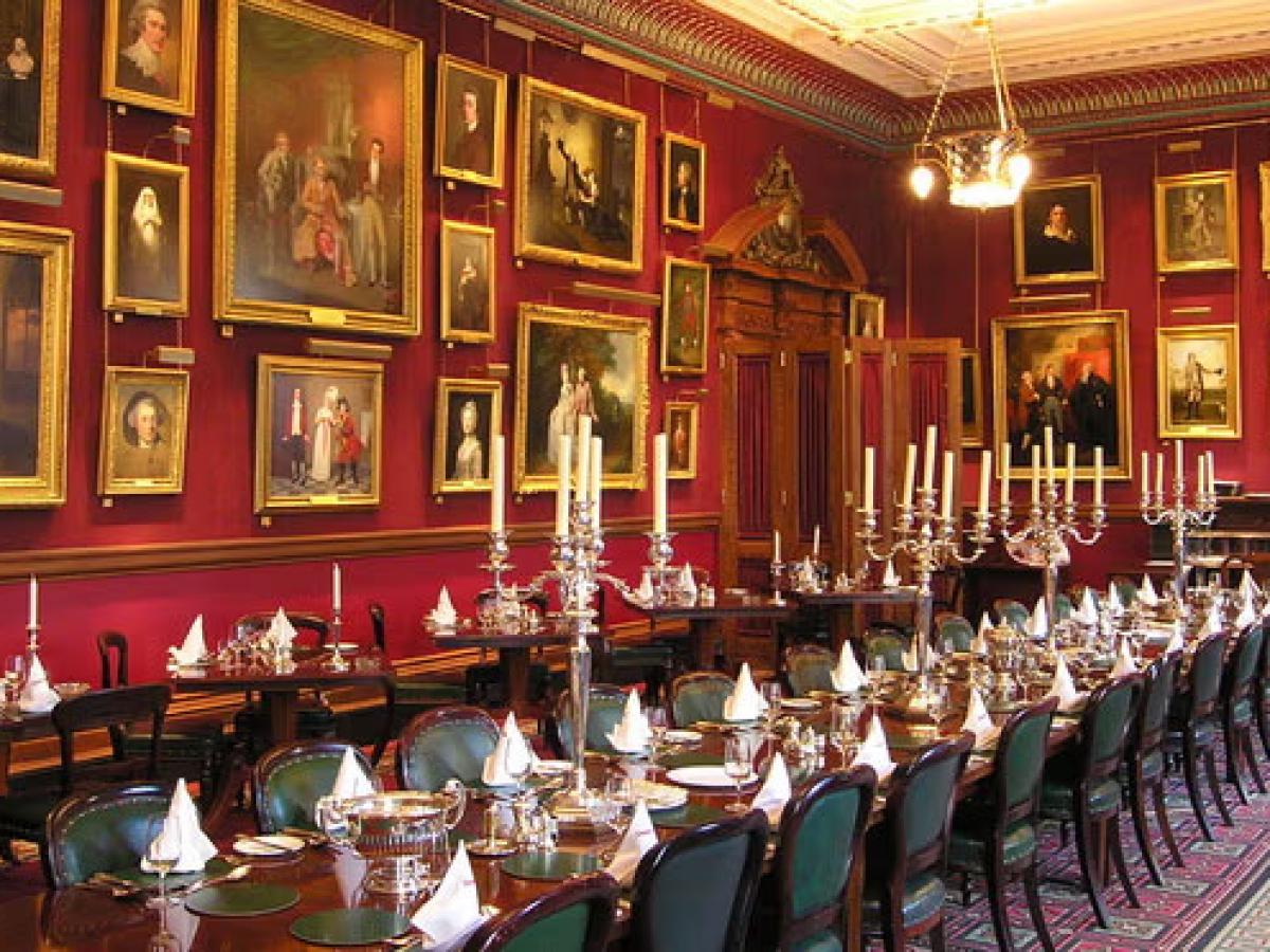 A grand dinner table set for a meal sits within a large room with paintings on the wall.