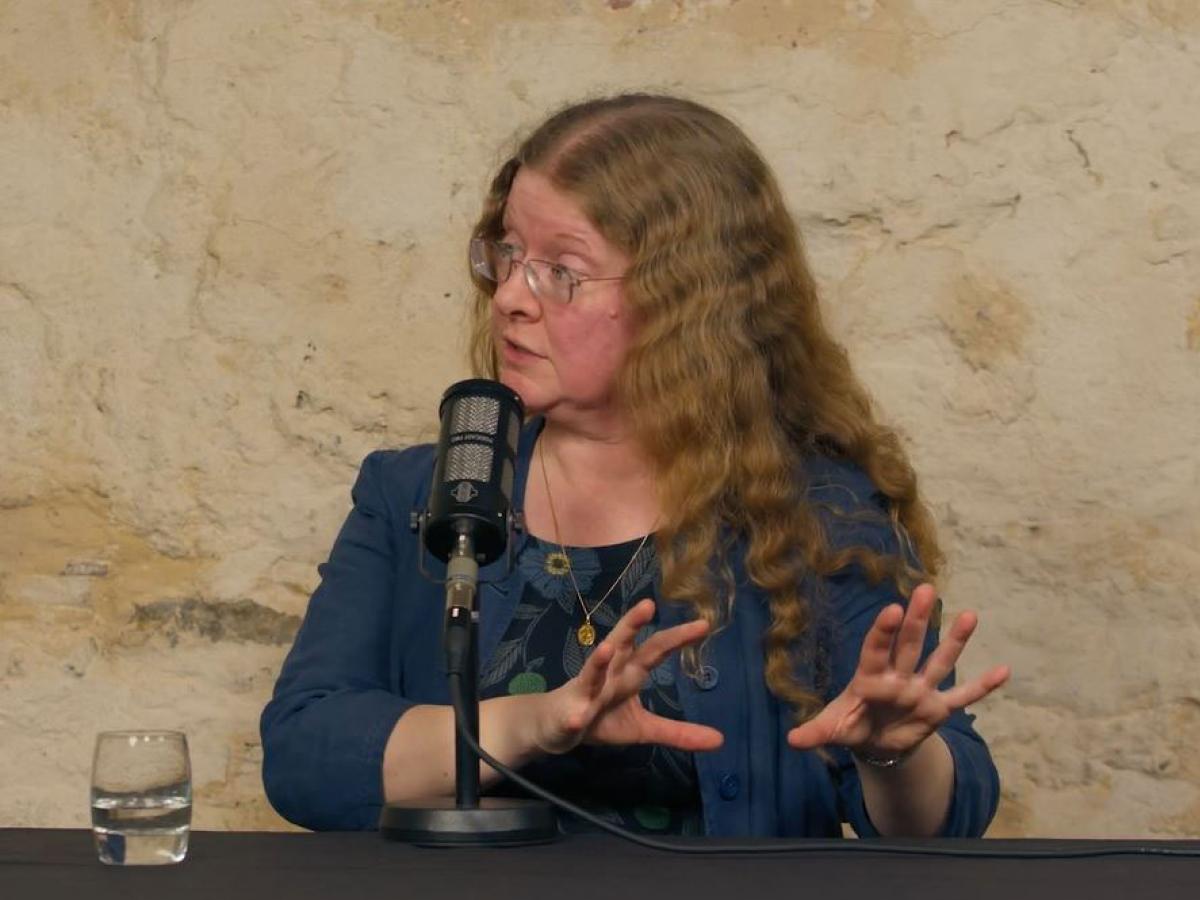 a woman seated in front a mic, looks to the left will gesturing both hands to the right.