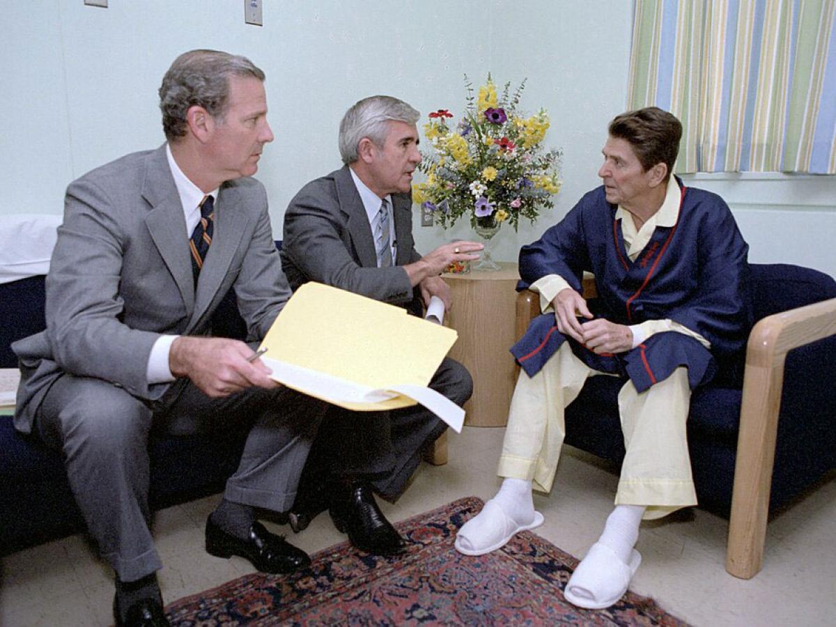 Two men in seats adress a pyjamap-clad Ronald Regan.