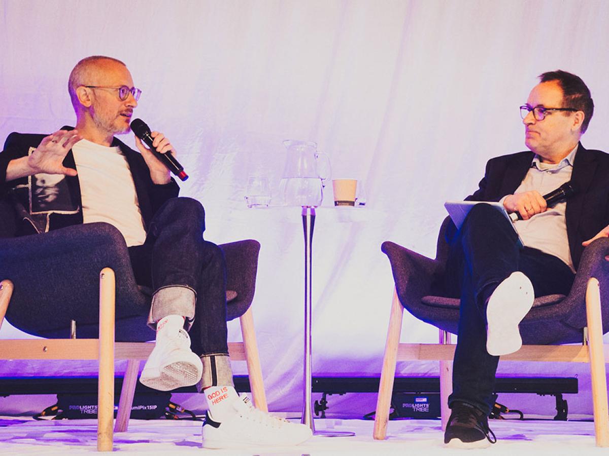 Two people sit on a stage in a relaxed manner, having a converation.