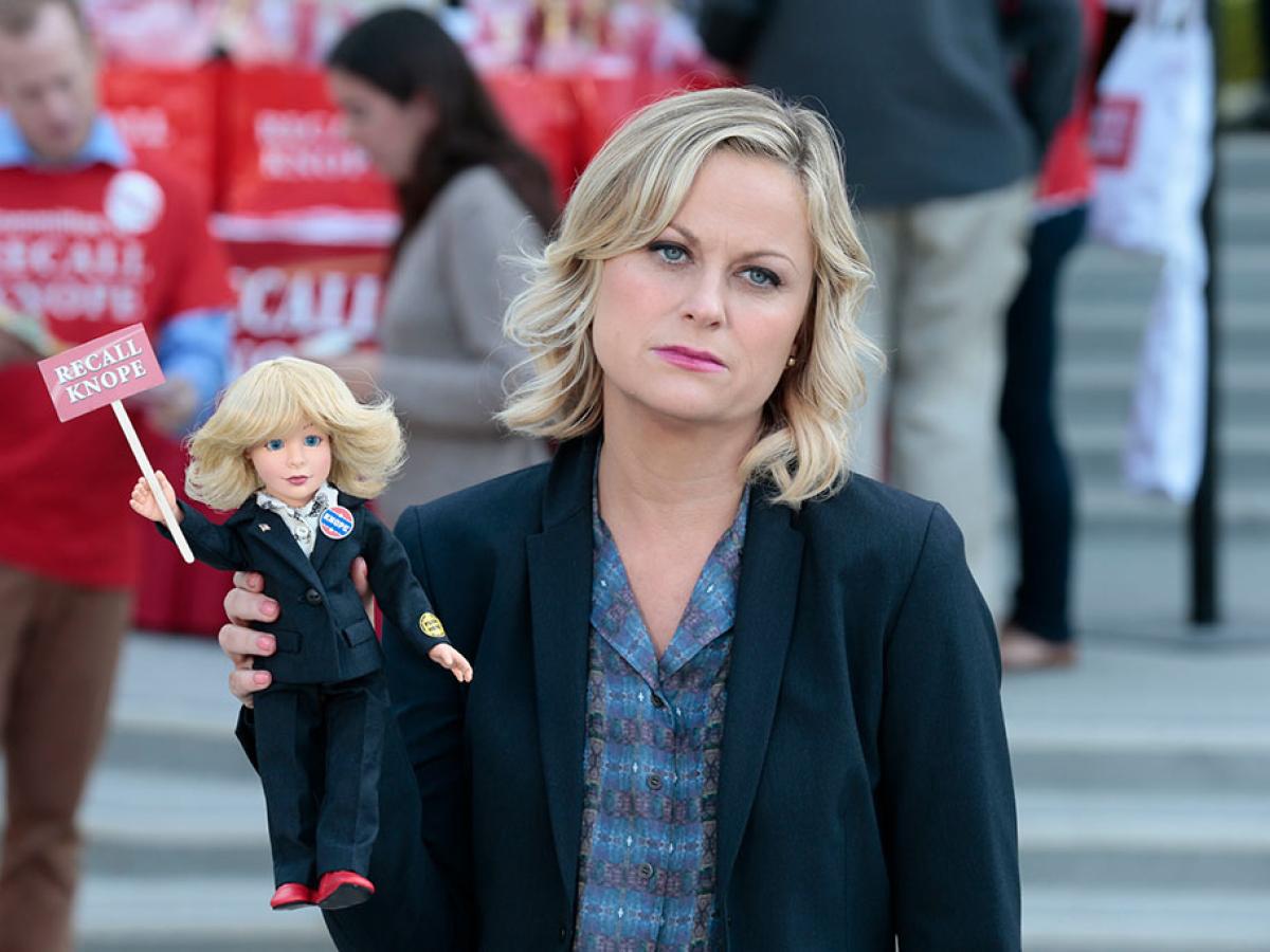 A deflated looking woman stands aside from a protest rally, holding a small doll of herself that reads Recall Knope'.