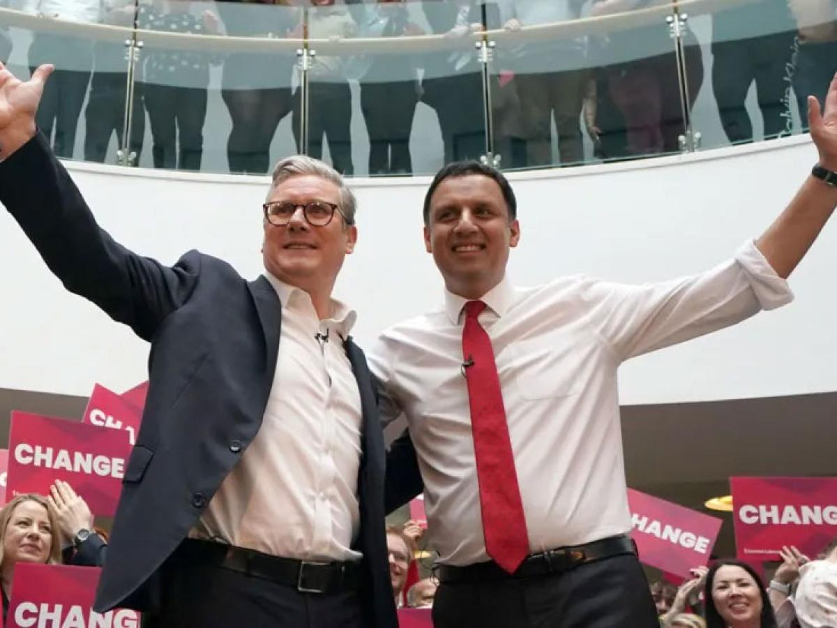 Two politicians, stand with one arm around each other and the other arms raised.