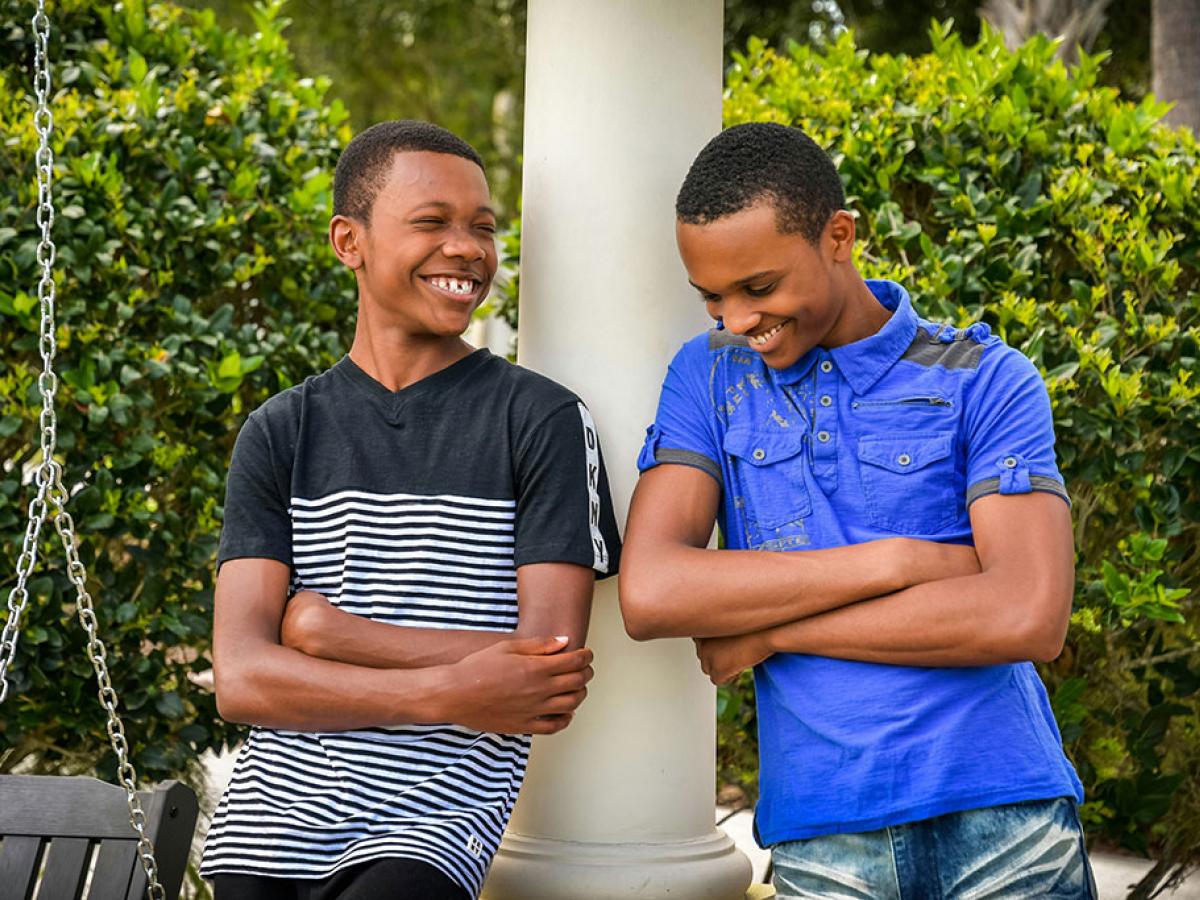 Two teenager lean against a rail, arms crossed, and laugh together.