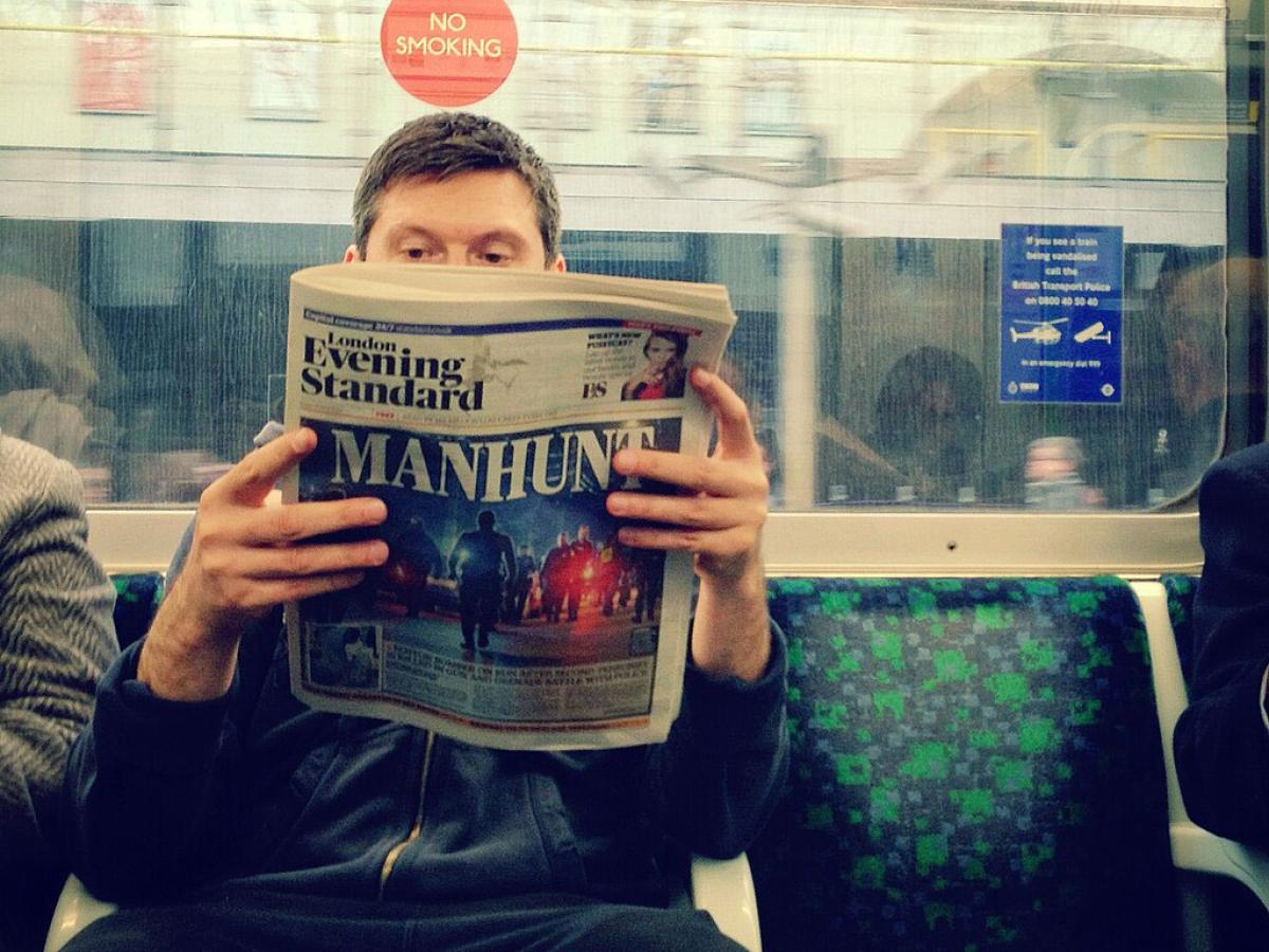 Seat on an underground train carraige, a passenger holds and reads a newspaper.