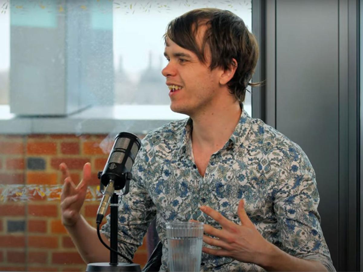 A podcast guest speaks in front of a mic and raises their hands to gesture.