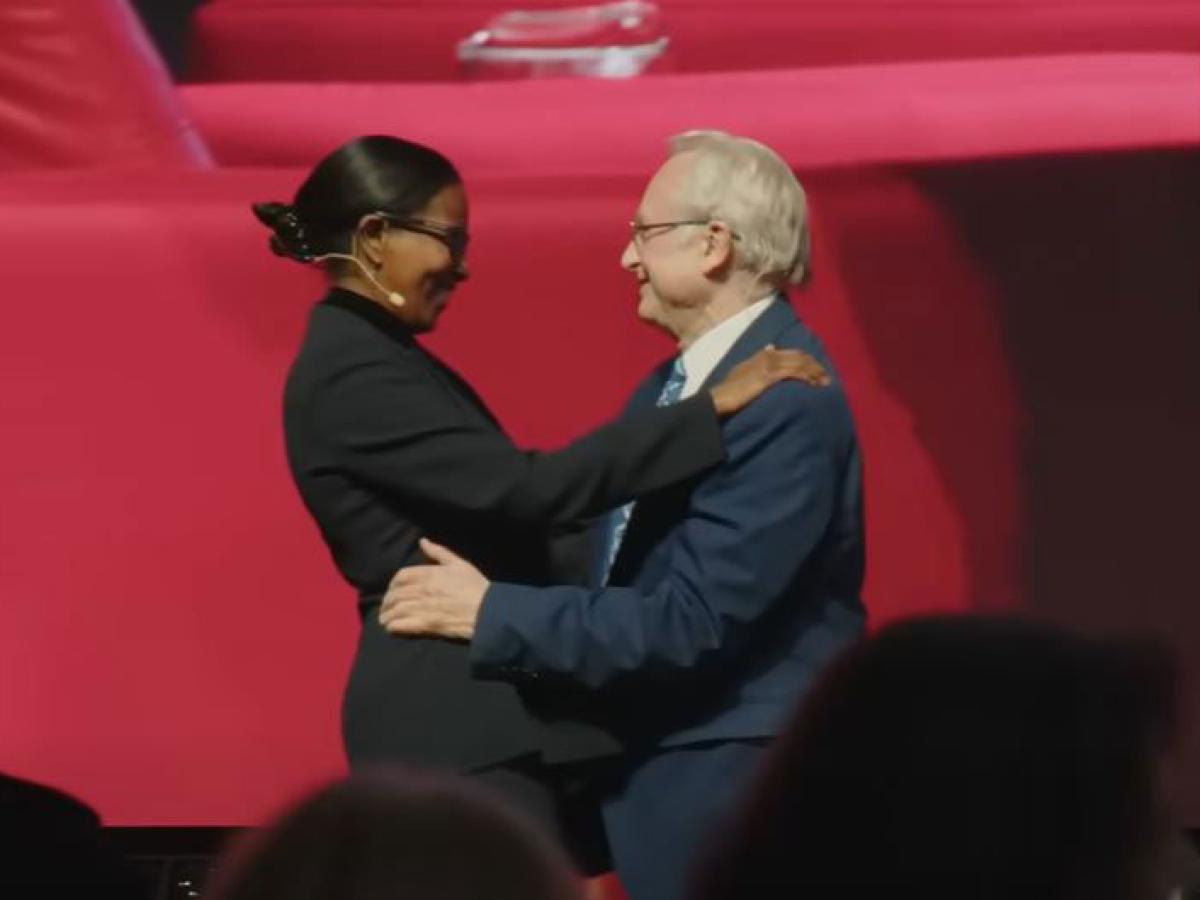 A man and woman speaker on a stage greet and embrace each other.