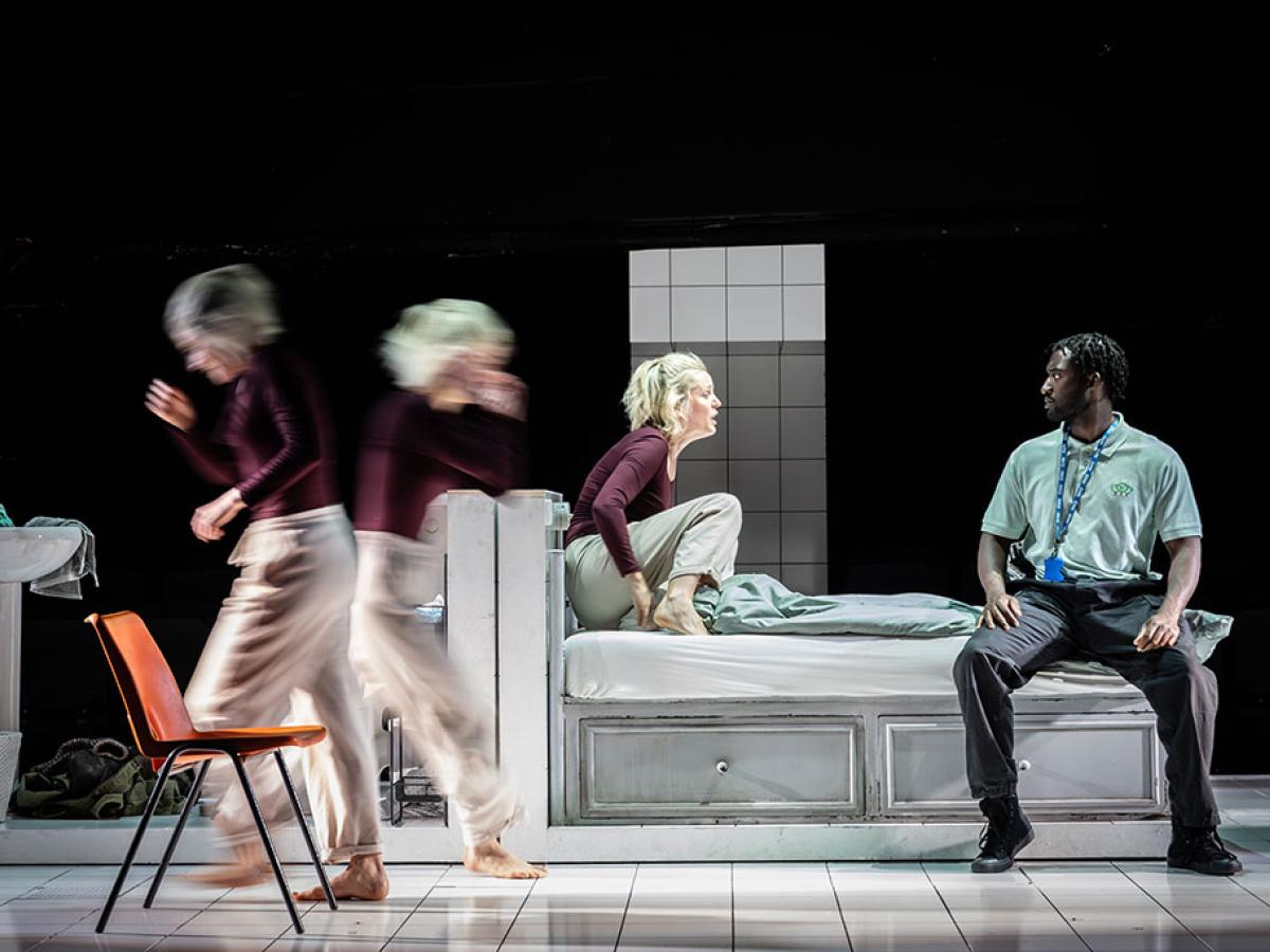 on a stage a woman kneels on a bed amid frantic action around her.