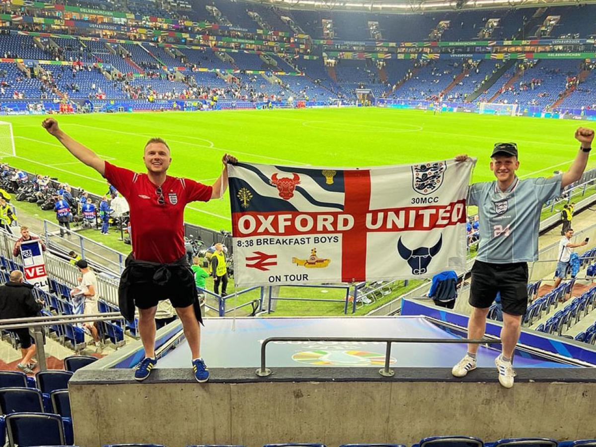 Two England fans stand in stadium seeting holding up their flag.