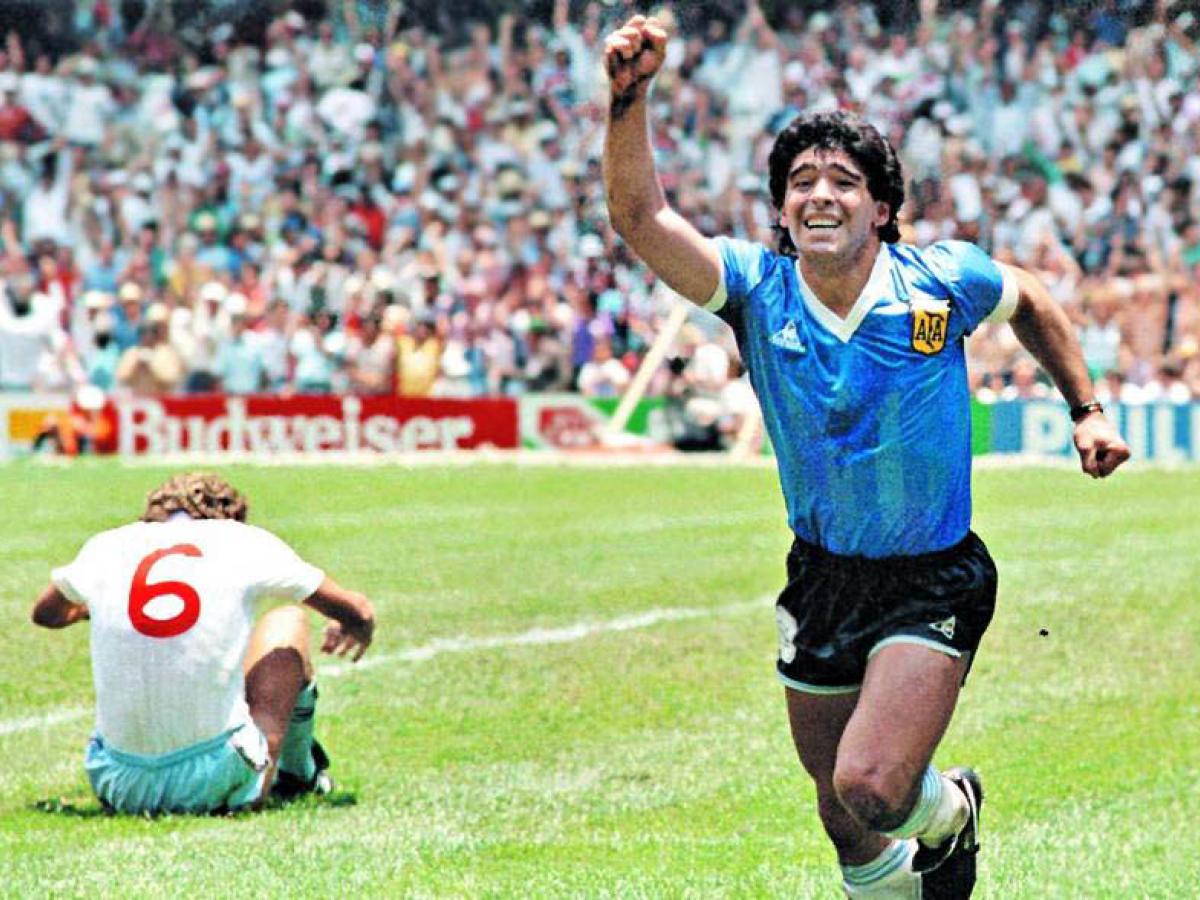 Maradona runs in celebration, holding a hand aloft as an England player sits dejected on the ground.