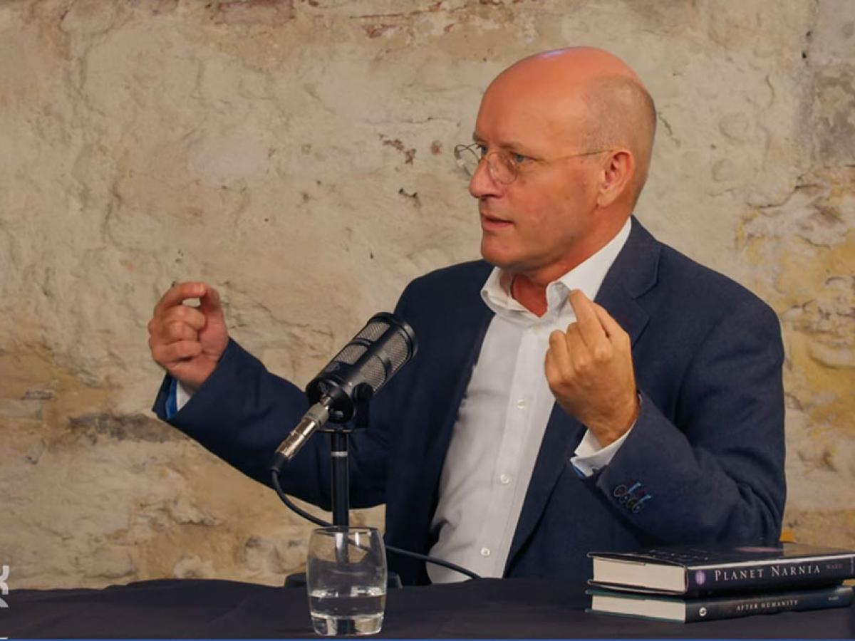 A man sits and speaks into a microphone and gestures with raised hands.