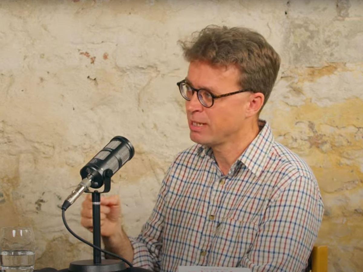 A podcast guest speaks in front of a mic and raises their hands to gesture.