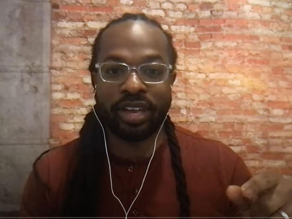 A man wearing white earphones talks to a camera and gestures with one hand.