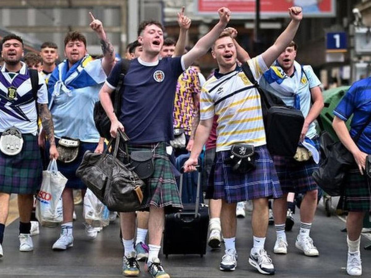 Scottish football fans wearing kilts march down a street singing and waving their arms alogt.